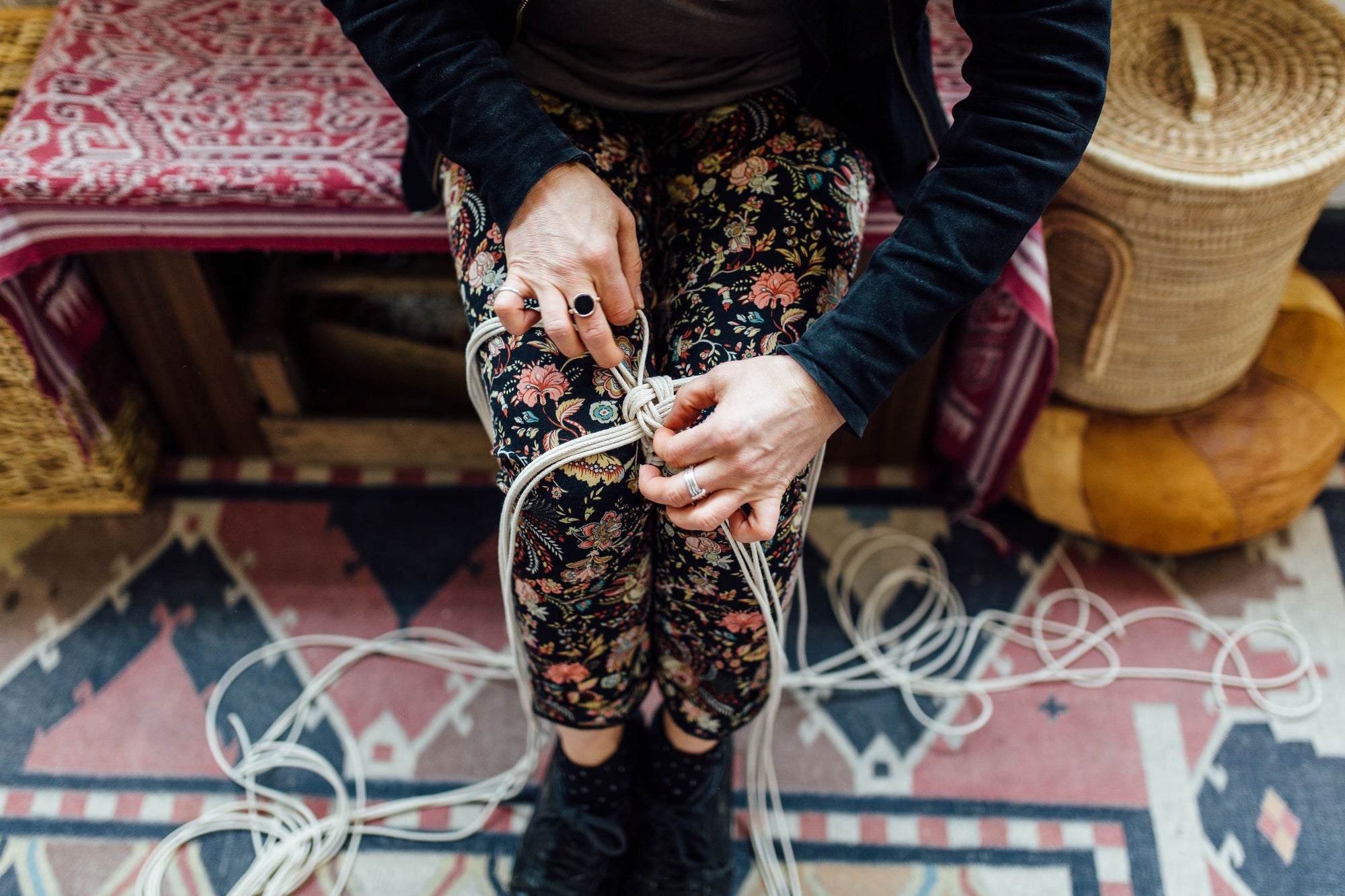 Person with a bundle of cords tying a knot over a patterned rug with a woven basket in the background